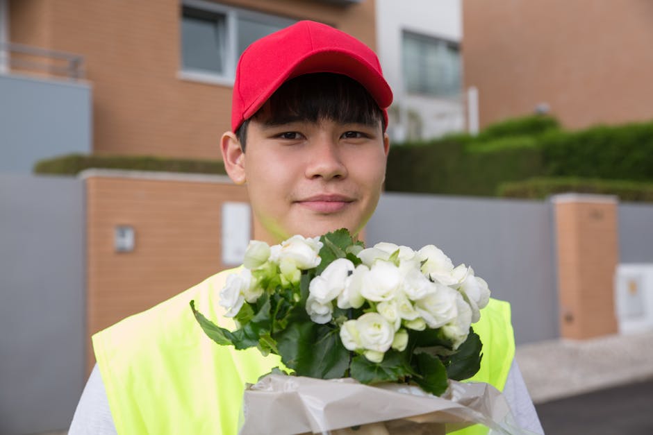 A smiling young delivery person wearing a red cap and a high-visibility yellow vest holds a bouquet of fresh white flowers, including roses and possibly freesias, wrapped in white paper. The flowers a