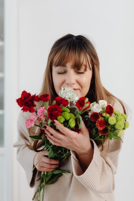 A young woman with light brown hair and bangs gently holding a vibrant bouquet of fresh flowers close to her face. The bouquet includes red carnations, pink roses, white hydrangeas, and small green bu