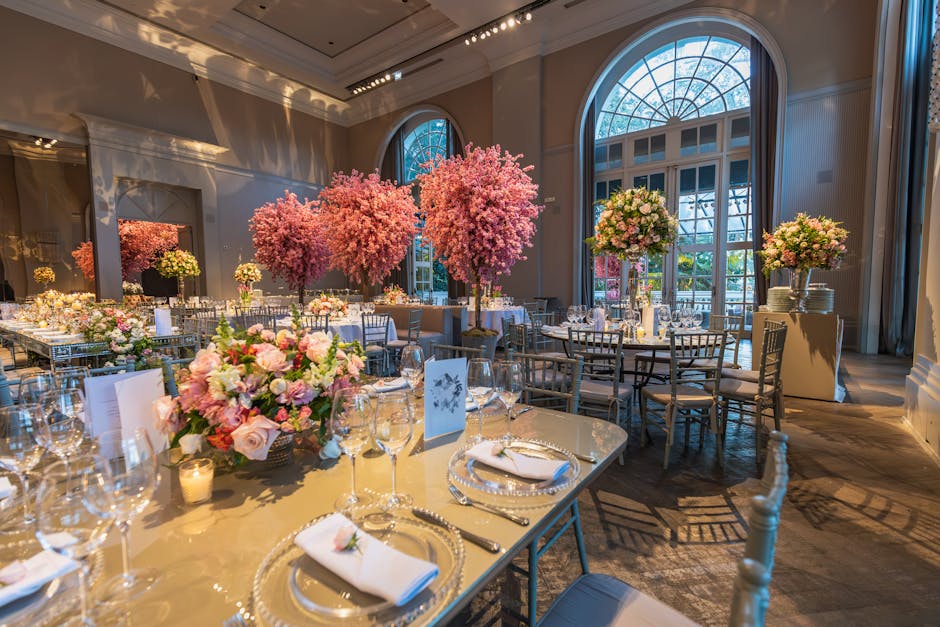 An elegant indoor event space featuring a sophisticated table setting with a lush floral arrangement centerpiece of pink roses, peonies, and greenery, arranged in a low, rounded bouquet. The tables ar