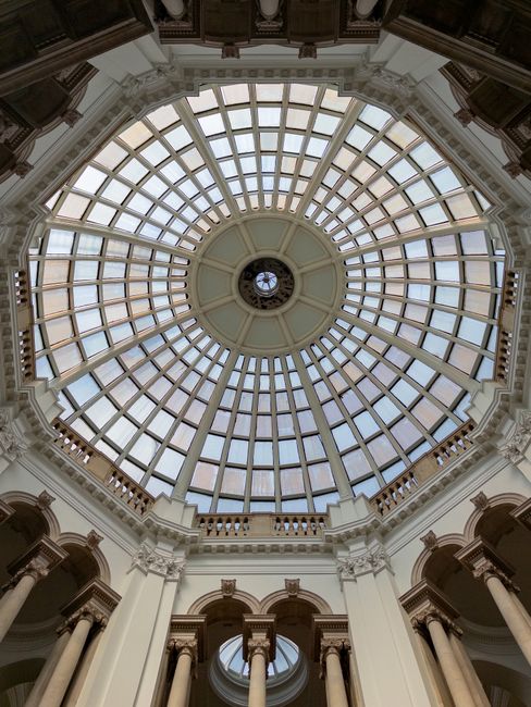 A detailed, symmetrical view of an ornate glass-domed ceiling inside a historic building, featuring a circular pattern with multiple rectangular glass panels bordered by white decorative molding. The 