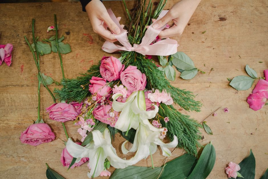 A pair of hands arranging a floral bouquet on a wooden surface, featuring pink roses, white lilies, and assorted greenery wrapped with pink ribbon. The bouquet includes fresh, vibrant flowers with lus