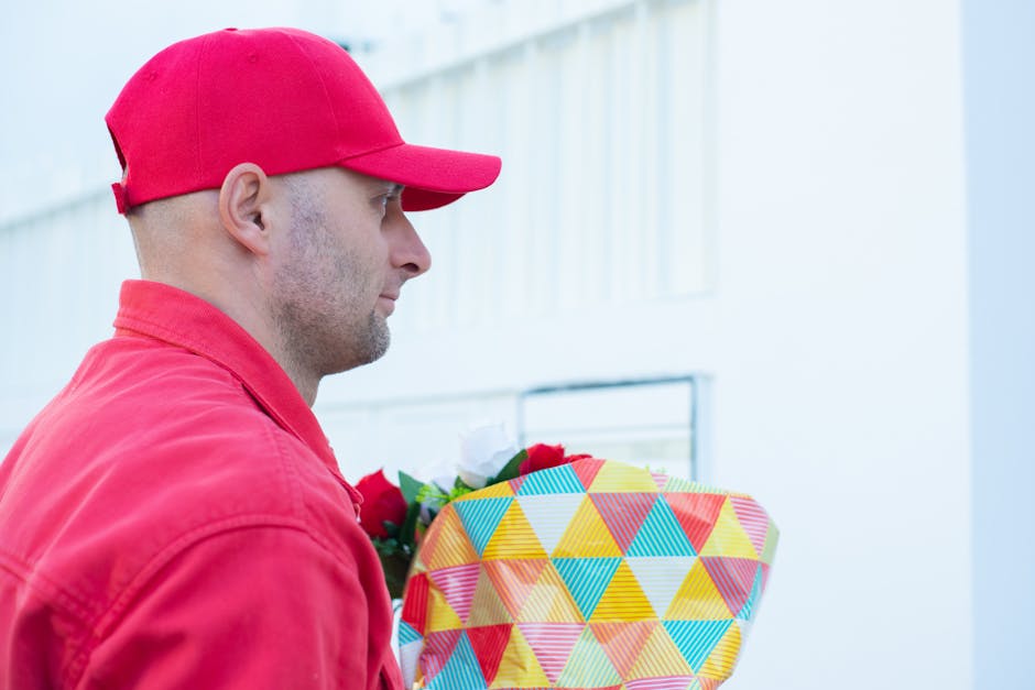 A man dressed in a red cap and jacket is holding a floral gift wrapped in colorful, geometric-patterned paper, featuring shades of yellow, orange, blue, and pink. The bouquet contains vibrant red and