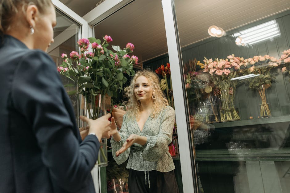 A woman with curly blonde hair, dressed in a floral patterned blouse, is receiving a bouquet of mixed pink and green flowers from a florist at a flower shop. The florist, visible from behind, is weari